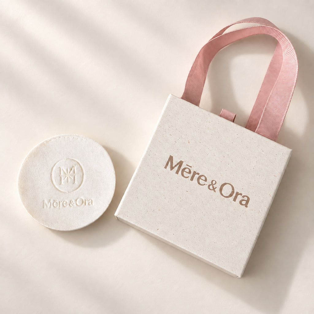 Mēre&Ora ivory jewelry box with a light brown logo, soft pink ribbon handles, and a round embossed brand insert.