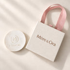 Mēre&Ora ivory jewelry box with a light brown logo, soft pink ribbon handles, and a round embossed brand insert.