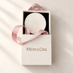 Partially open Mēre&Ora jewelry box with a black interior, round ivory embossed insert, and soft pink ribbon handles.