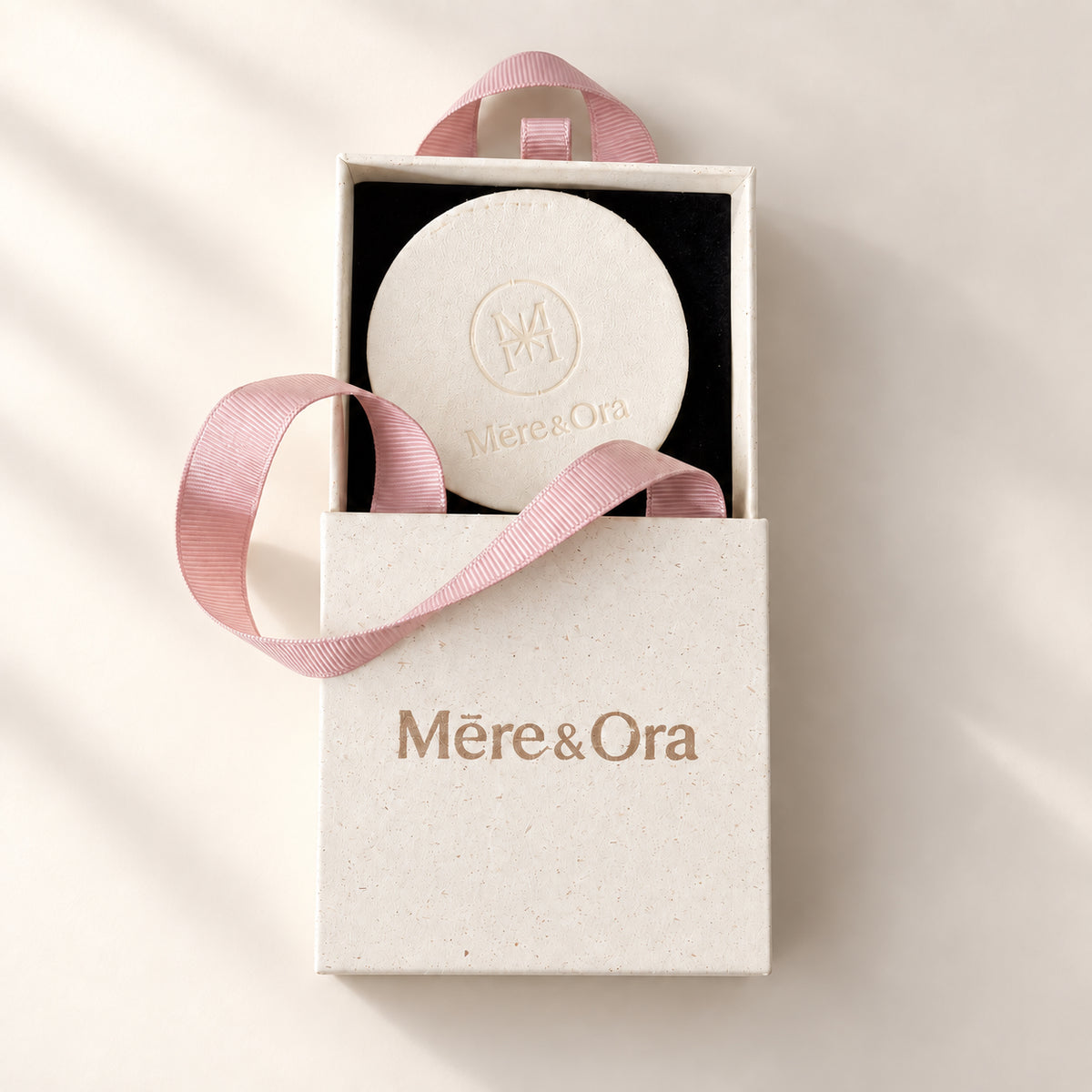 Partially open Mēre&Ora jewelry box with a black interior, round ivory embossed insert, and soft pink ribbon handles.