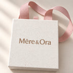 Close-up of Mēre&Ora packaging showing textured ivory paper, a light brown logo, and soft pink grosgrain ribbon handles.