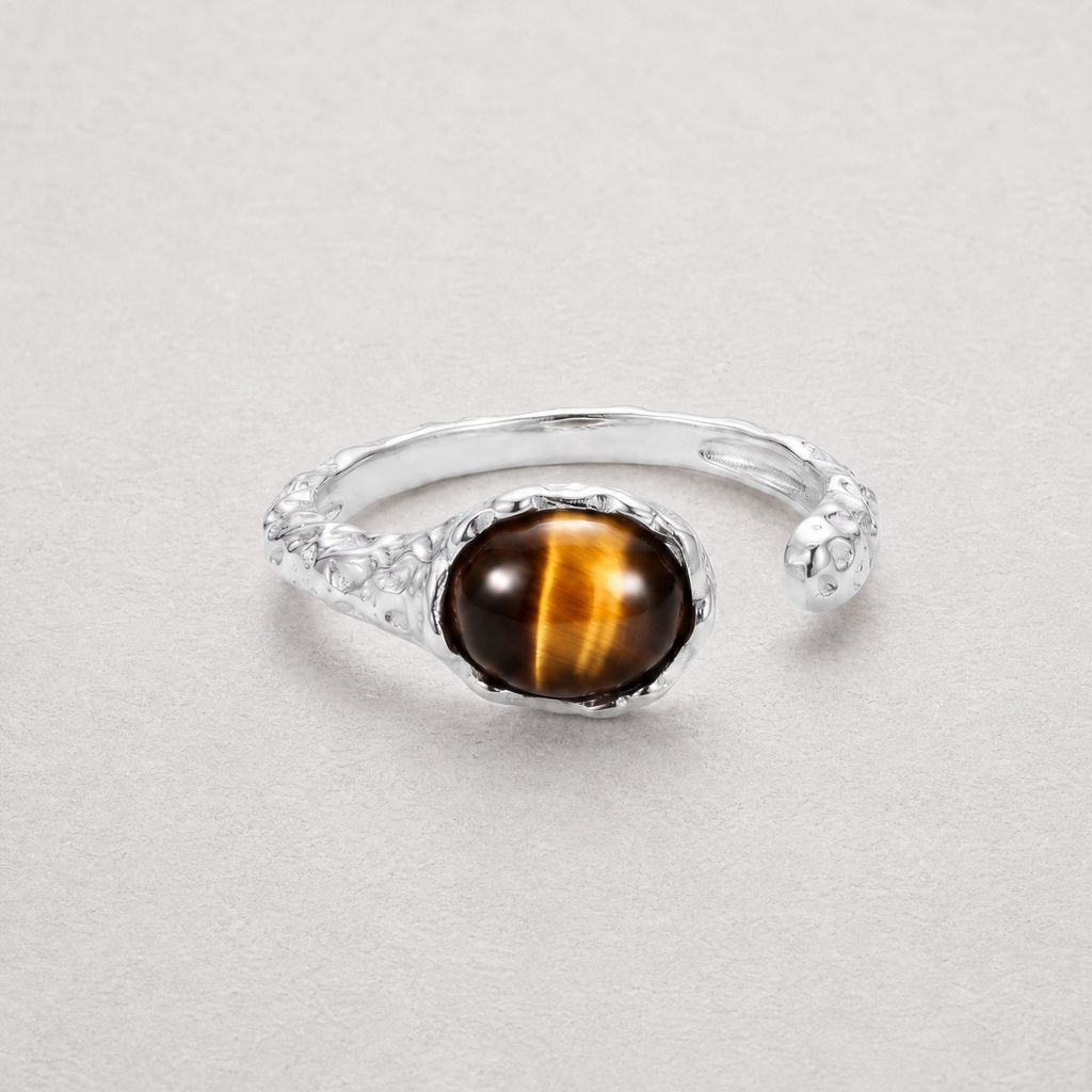A silver-toned open band ring featuring a round cabochon tiger's eye gemstone with its characteristic brown and gold stripes, set against a neutral light gray background.