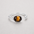 A silver-toned open band ring featuring a round cabochon tiger's eye gemstone with its characteristic brown and gold stripes, set against a neutral light gray background.