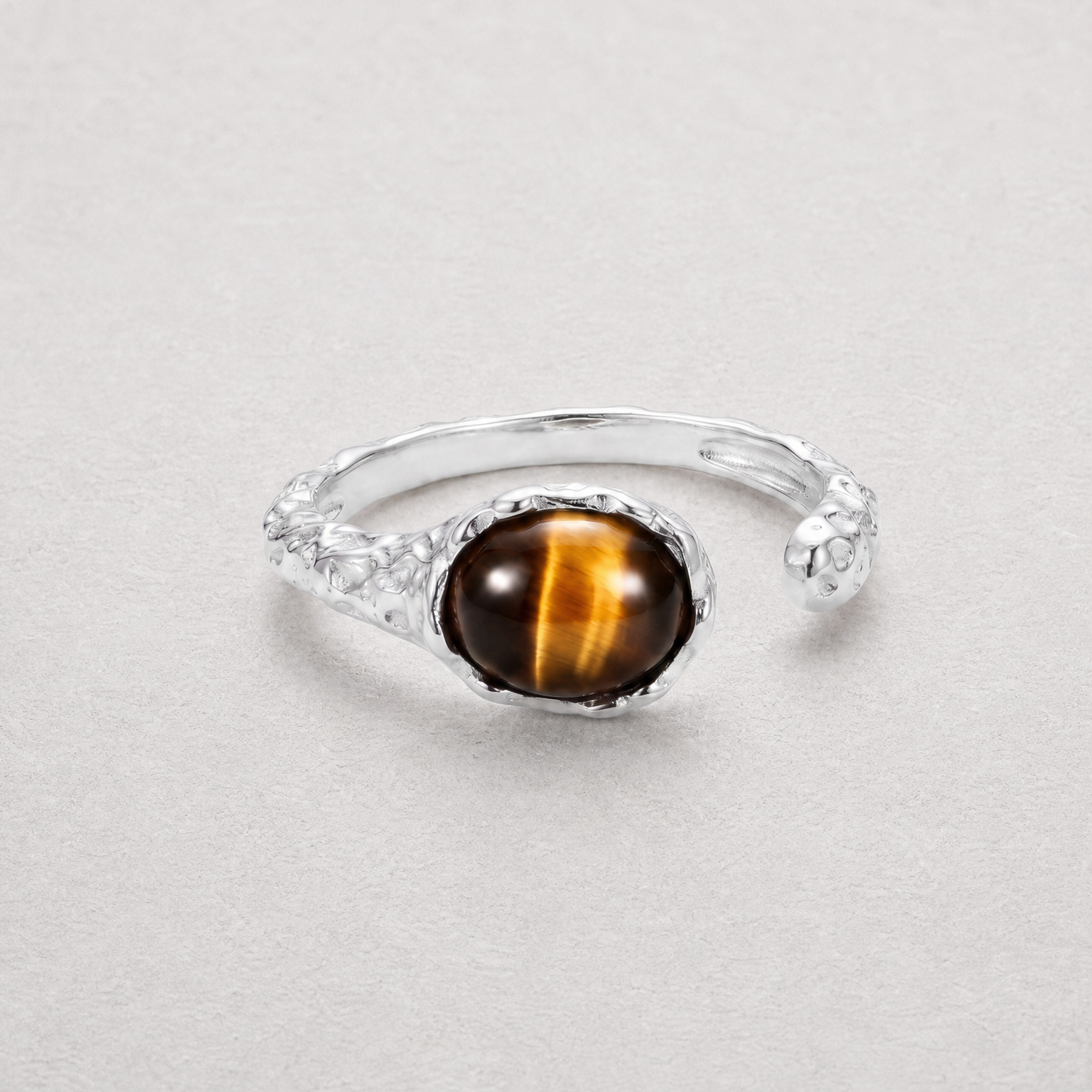 A silver-toned open band ring featuring a round cabochon tiger's eye gemstone with its characteristic brown and gold stripes, set against a neutral light gray background.