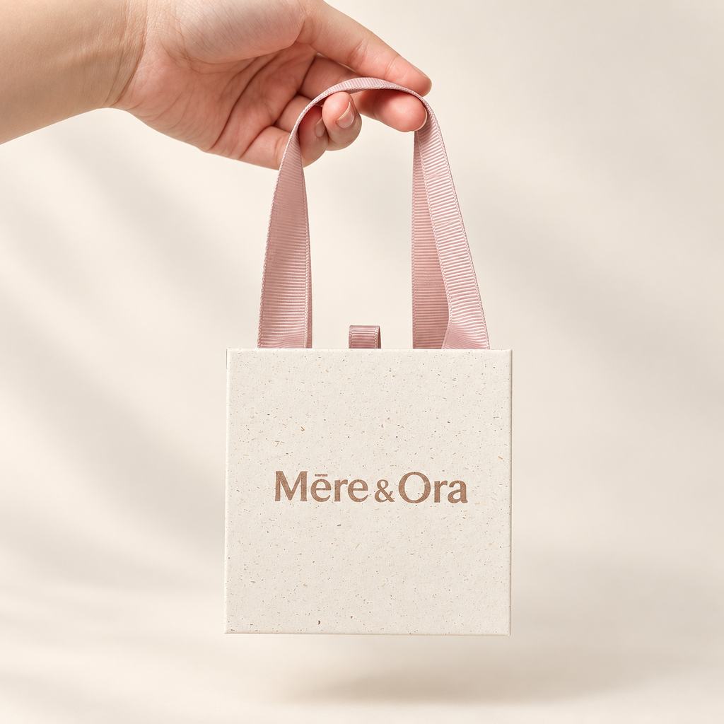 Hand holding a Mēre&Ora ivory jewelry box by soft pink ribbon handles, with a light brown logo against a warm white background.