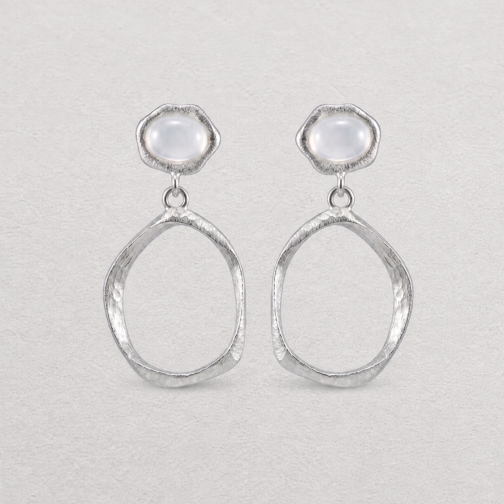 A pair of silver dangle earrings with textured oval drop shapes and smooth, dome-shaped white agate stones at the top, displayed on a light textured background.