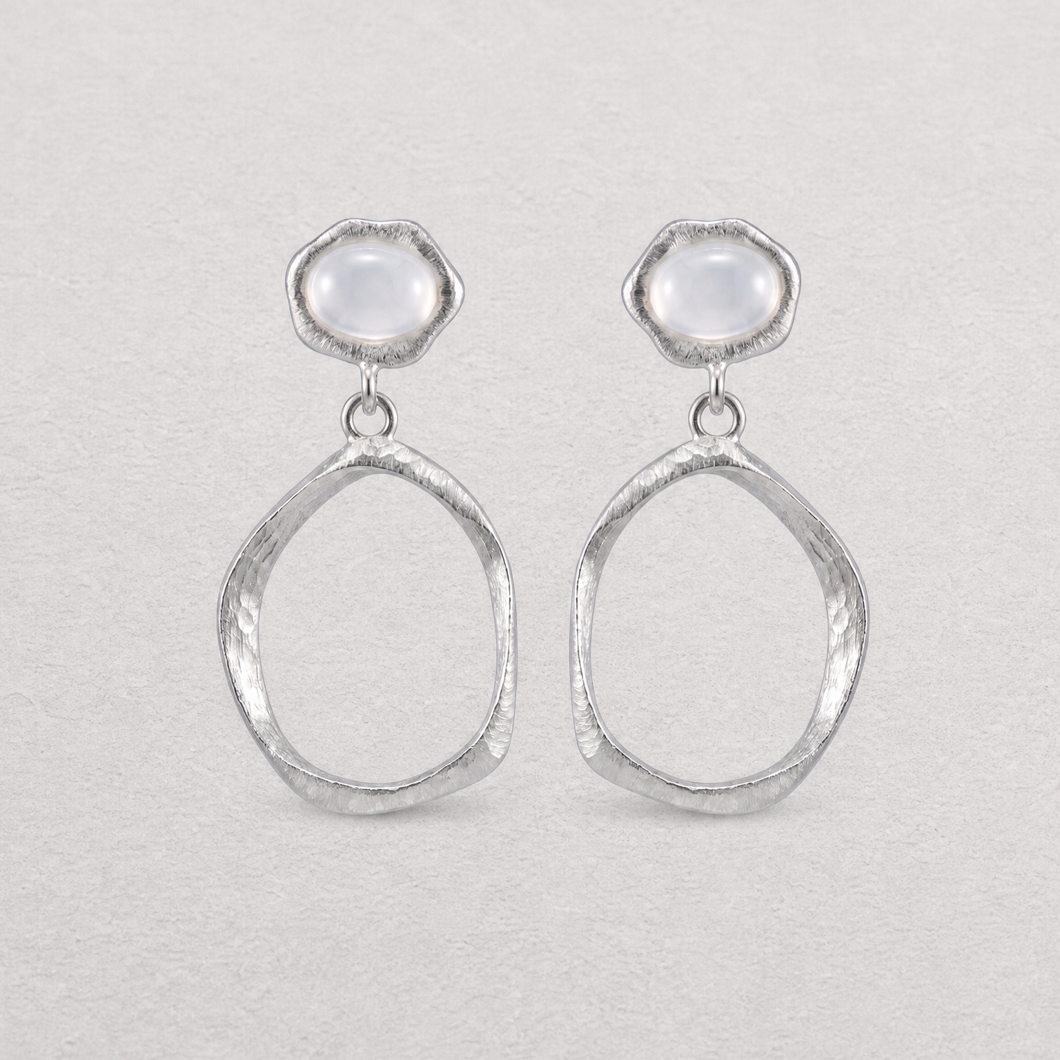 A pair of silver dangle earrings with textured oval drop shapes and smooth, dome-shaped white agate stones at the top, displayed on a light textured background.
