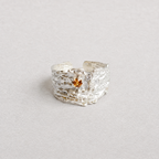 A textured silver ring with a small amber-colored gemstone in the center, photographed on a neutral light gray background.