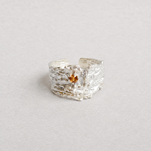 A textured silver ring with a small amber-colored gemstone in the center, photographed on a neutral light gray background.