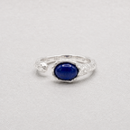 A sterling silver ring featuring a central oval cabochon lapis lazuli stone, photographed on a neutral light gray background.