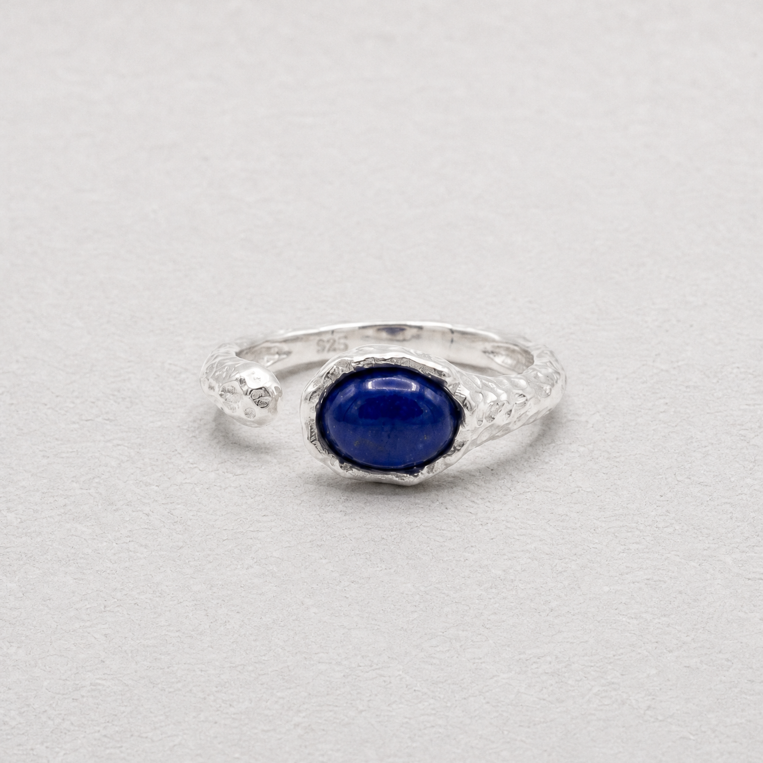 A sterling silver ring featuring a central oval cabochon lapis lazuli stone, photographed on a neutral light gray background.
