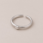 A minimalist silver open ring band featuring a small, polished heart charm on the front, set against a solid, warm beige background.