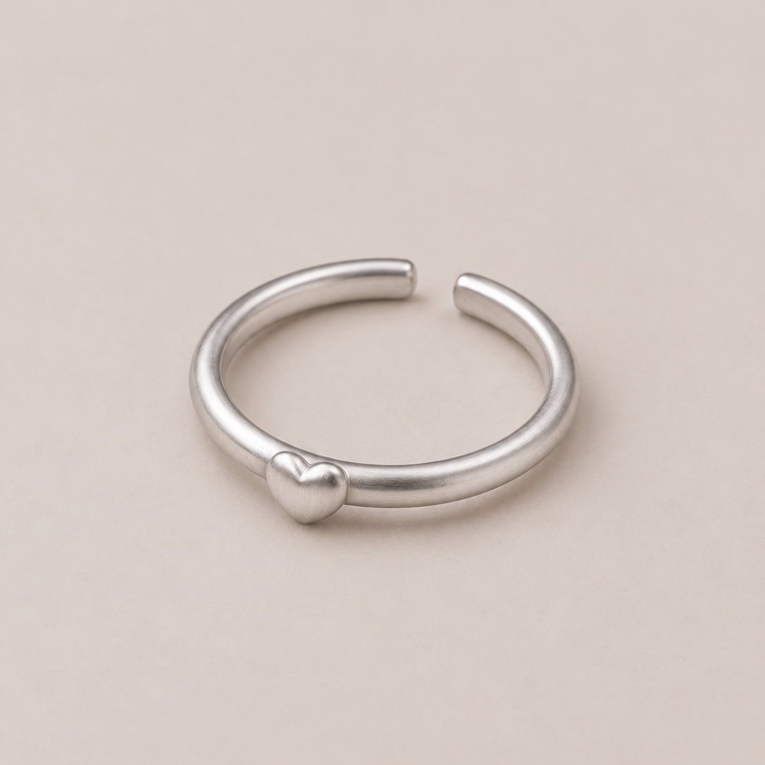 A minimalist silver open ring band featuring a small, polished heart charm on the front, set against a solid, warm beige background.