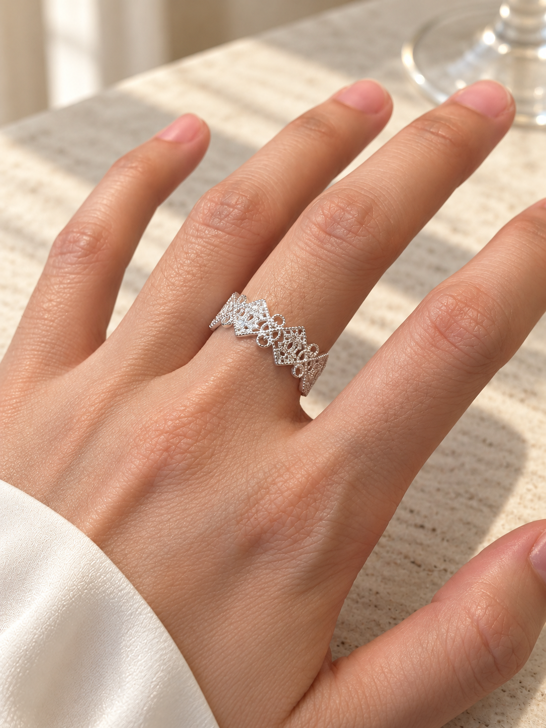 A close-up of a hand wearing a delicate silver ring with an intricate openwork floral or lace-like pattern, resting on a textured light beige surface.