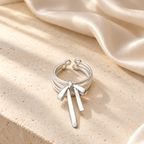 A silver-toned multi-band ring with a bow-shaped dangling charm, styled on a beige stone surface with draped satin fabric in soft sunlight.