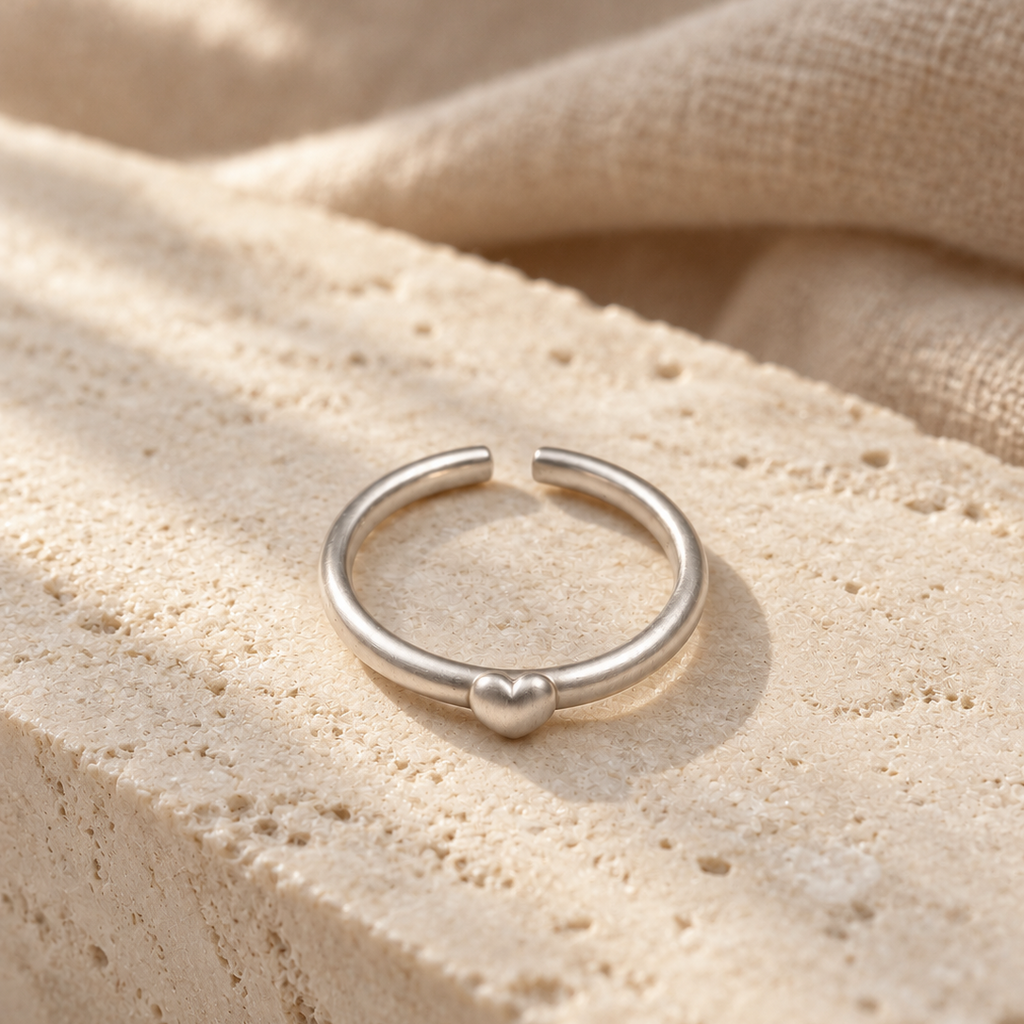 A minimalist silver ring with a small heart detail at the center, displayed on a textured beige stone surface with soft linen fabric in the background.