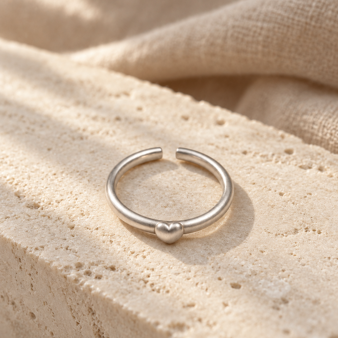 A minimalist silver ring with a small heart detail at the center, displayed on a textured beige stone surface with soft linen fabric in the background.