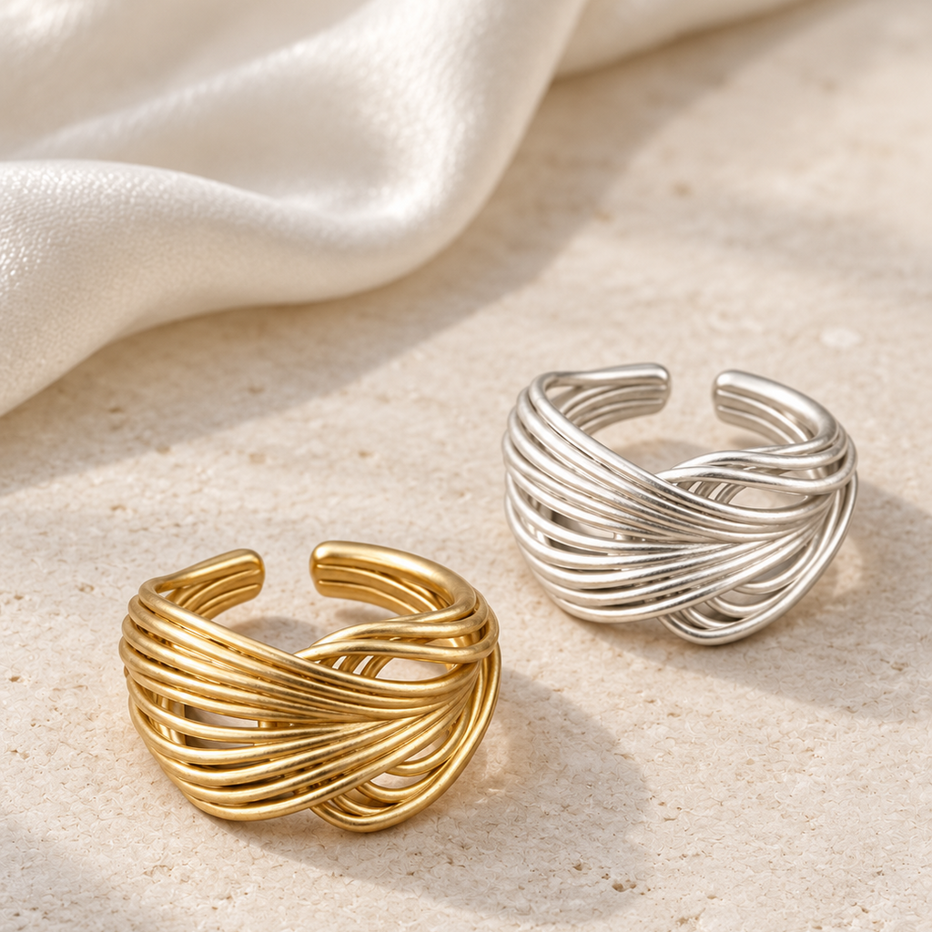 Two twisted open cuff rings, one in polished gold and one in polished silver, displayed on a textured beige stone surface with a draped white satin fabric in the background.