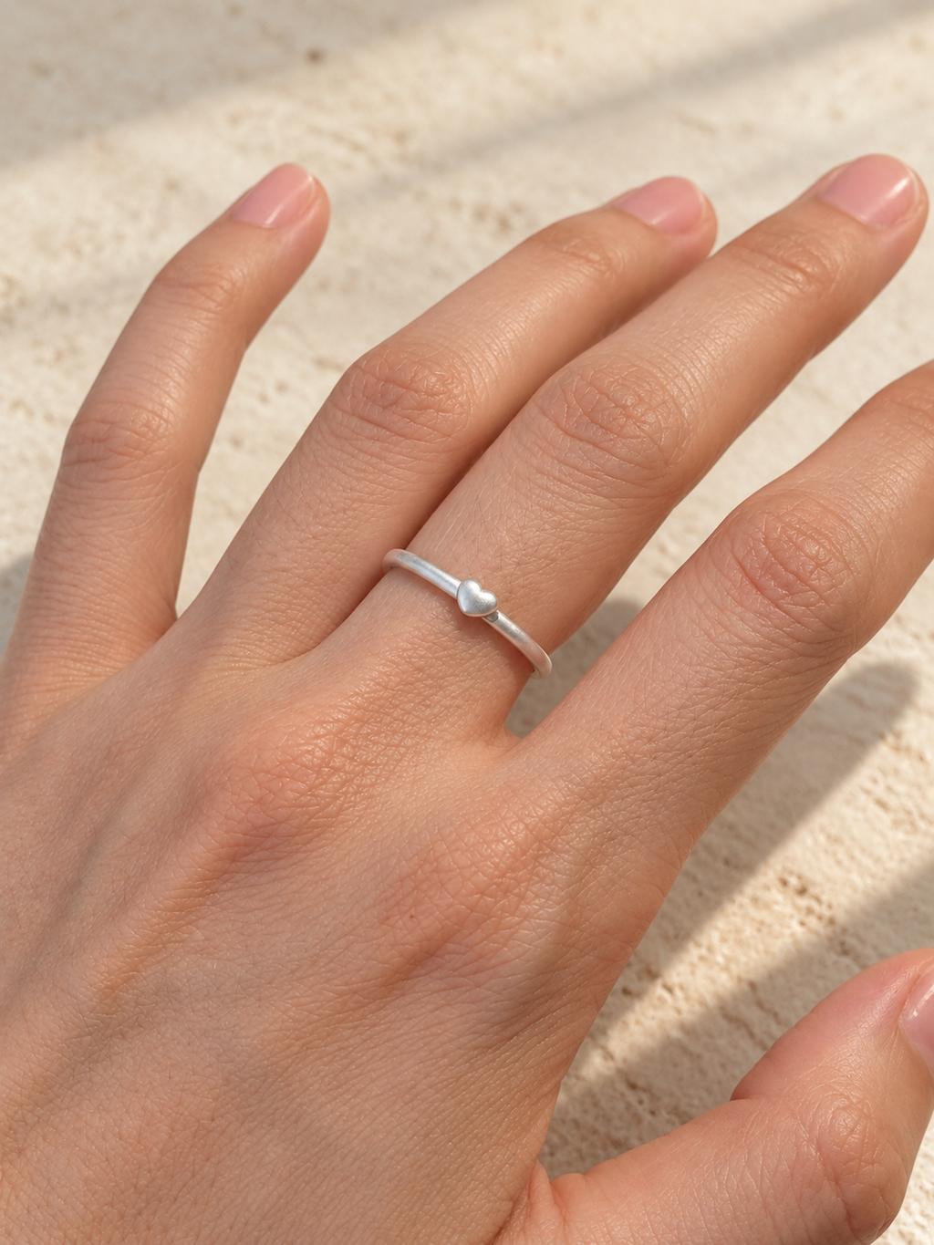 A close-up of a hand wearing a delicate silver band ring with a small heart-shaped detail, set against a textured beige background.