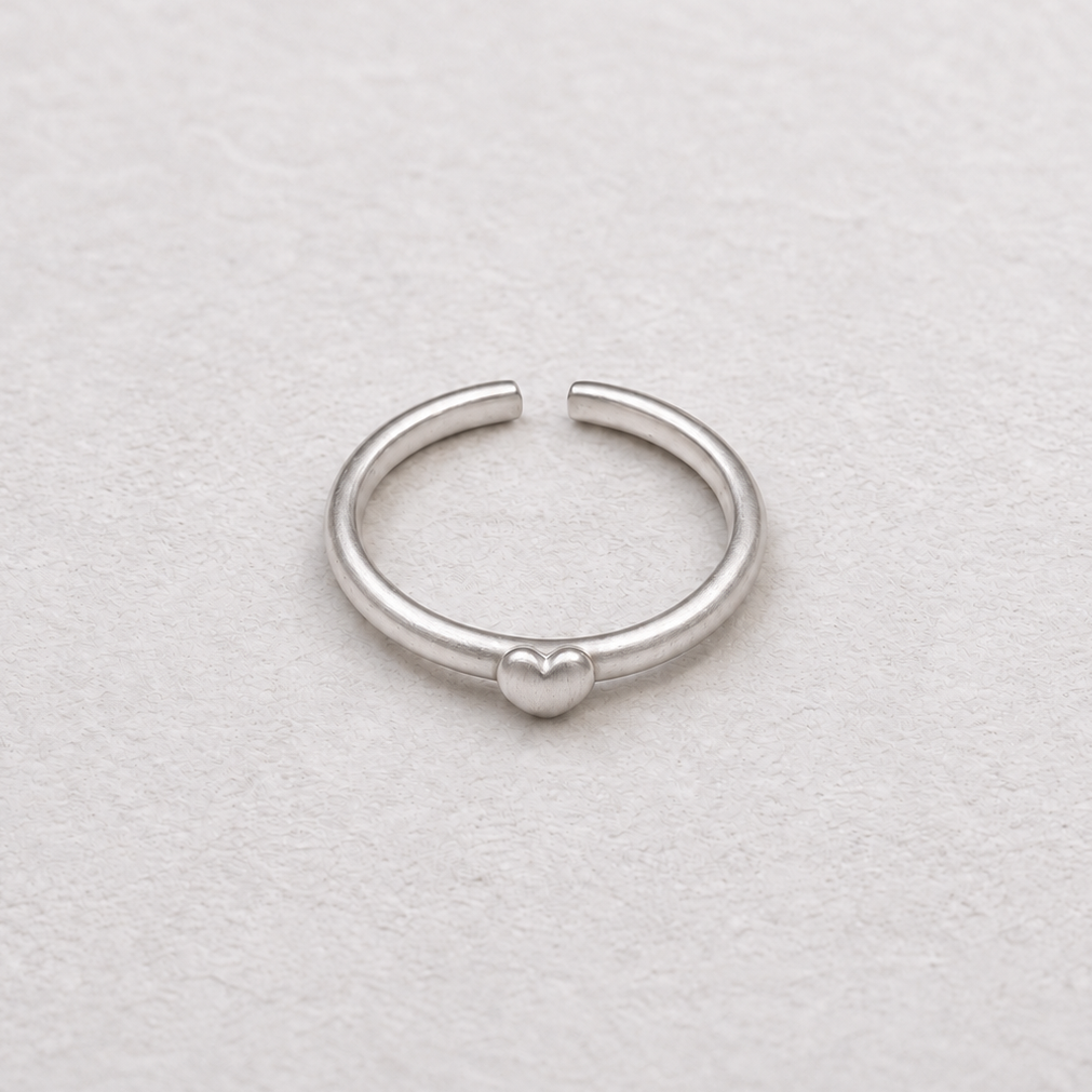 A minimalist silver ring with a small heart charm in the center, displayed on a textured off-white surface.