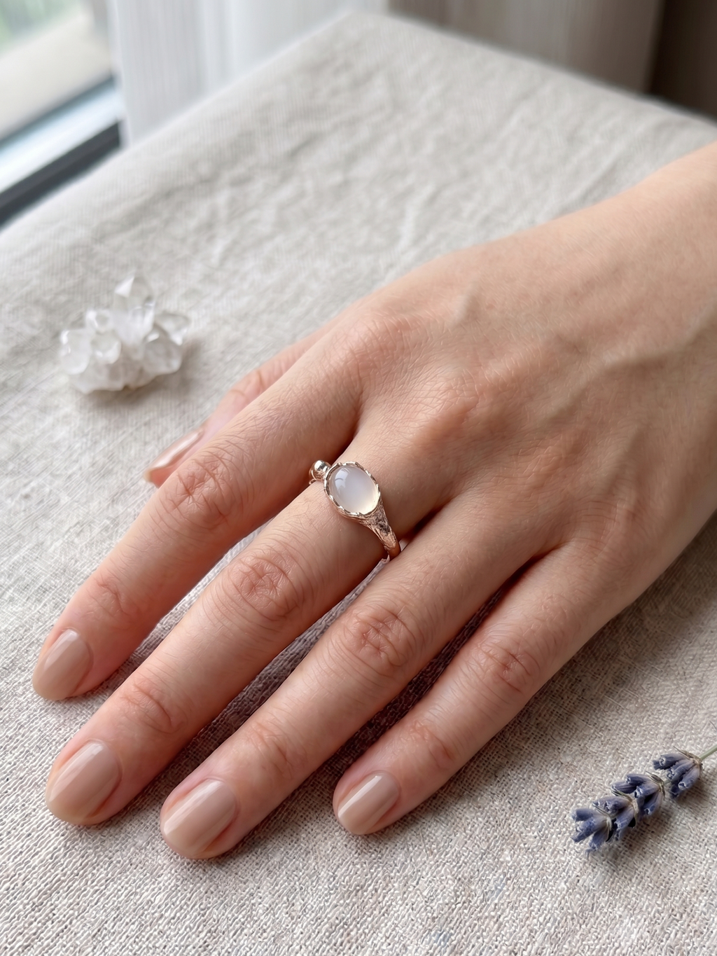 Hand wearing a white agate ring with a clear stone on a textured surface with lavender flowers.