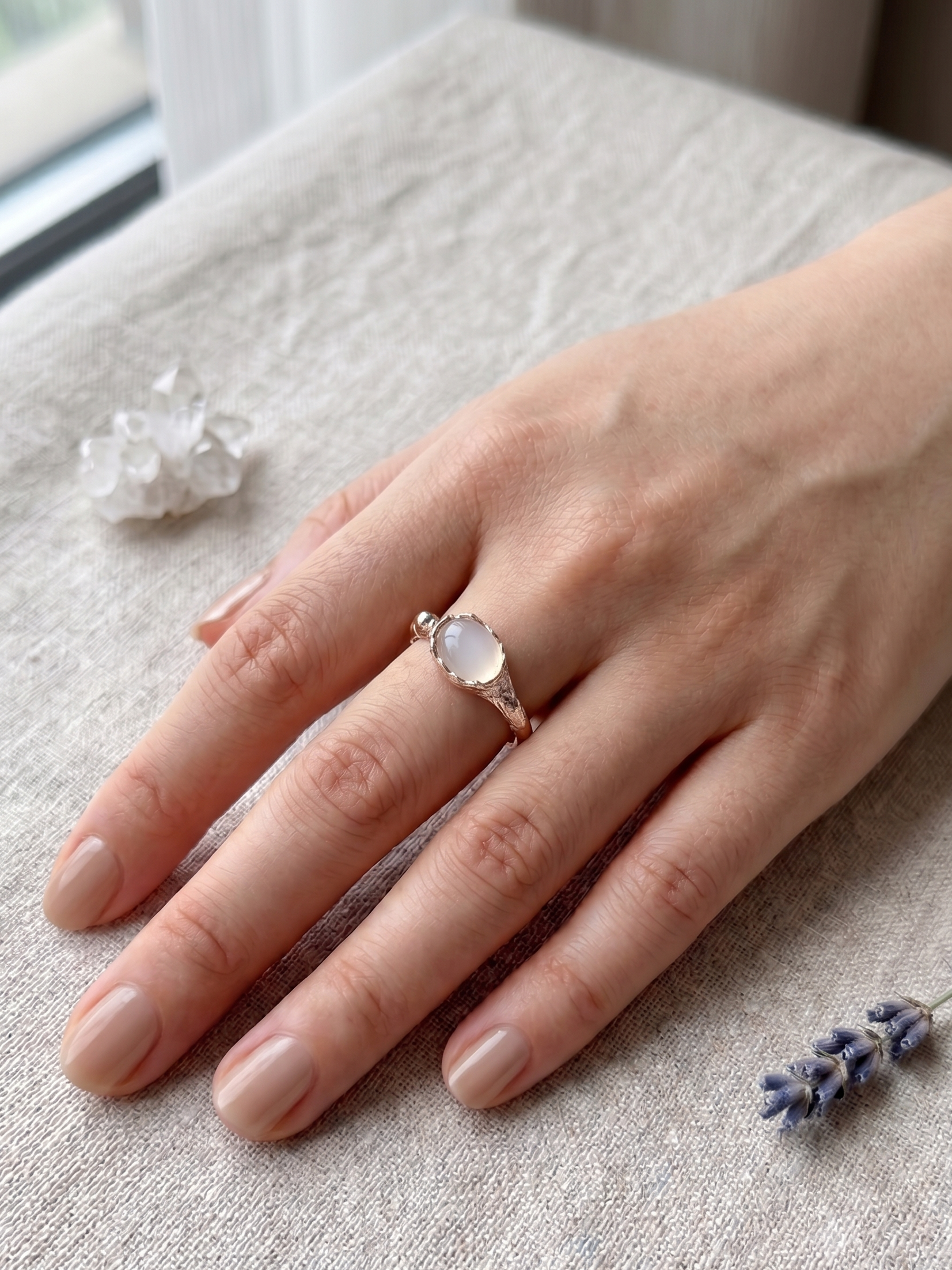 Hand wearing a white agate ring with a clear stone on a textured surface with lavender flowers.