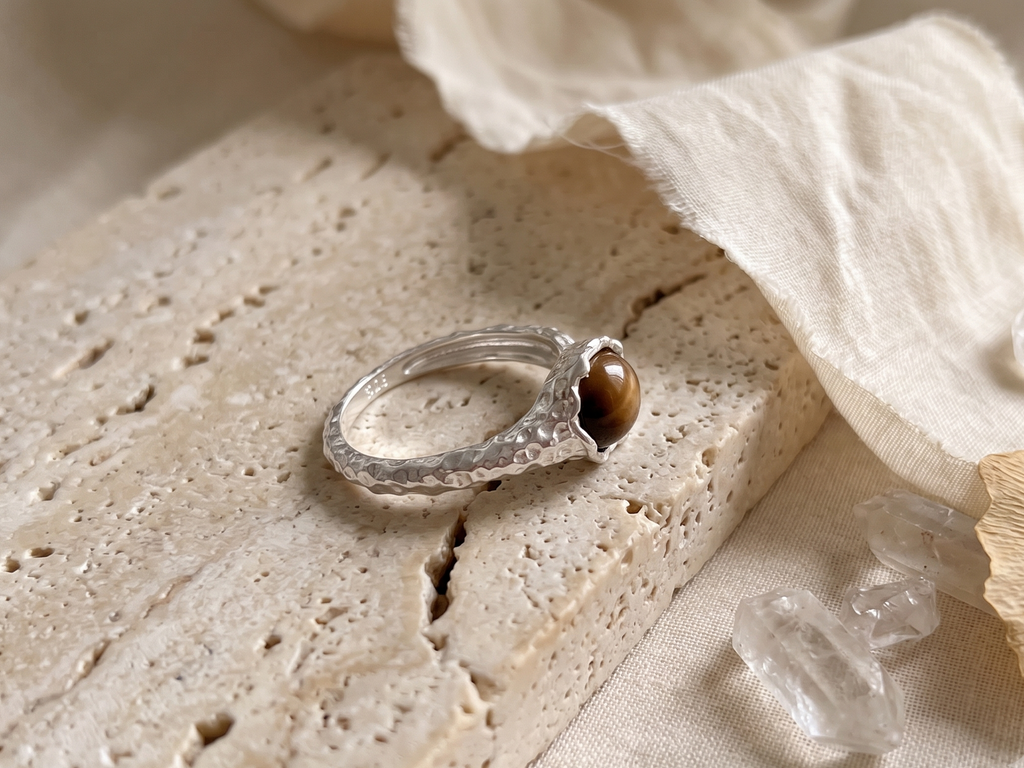 Silver ring with a tiger's eye stone on a textured beige surface with crystals and fabric.