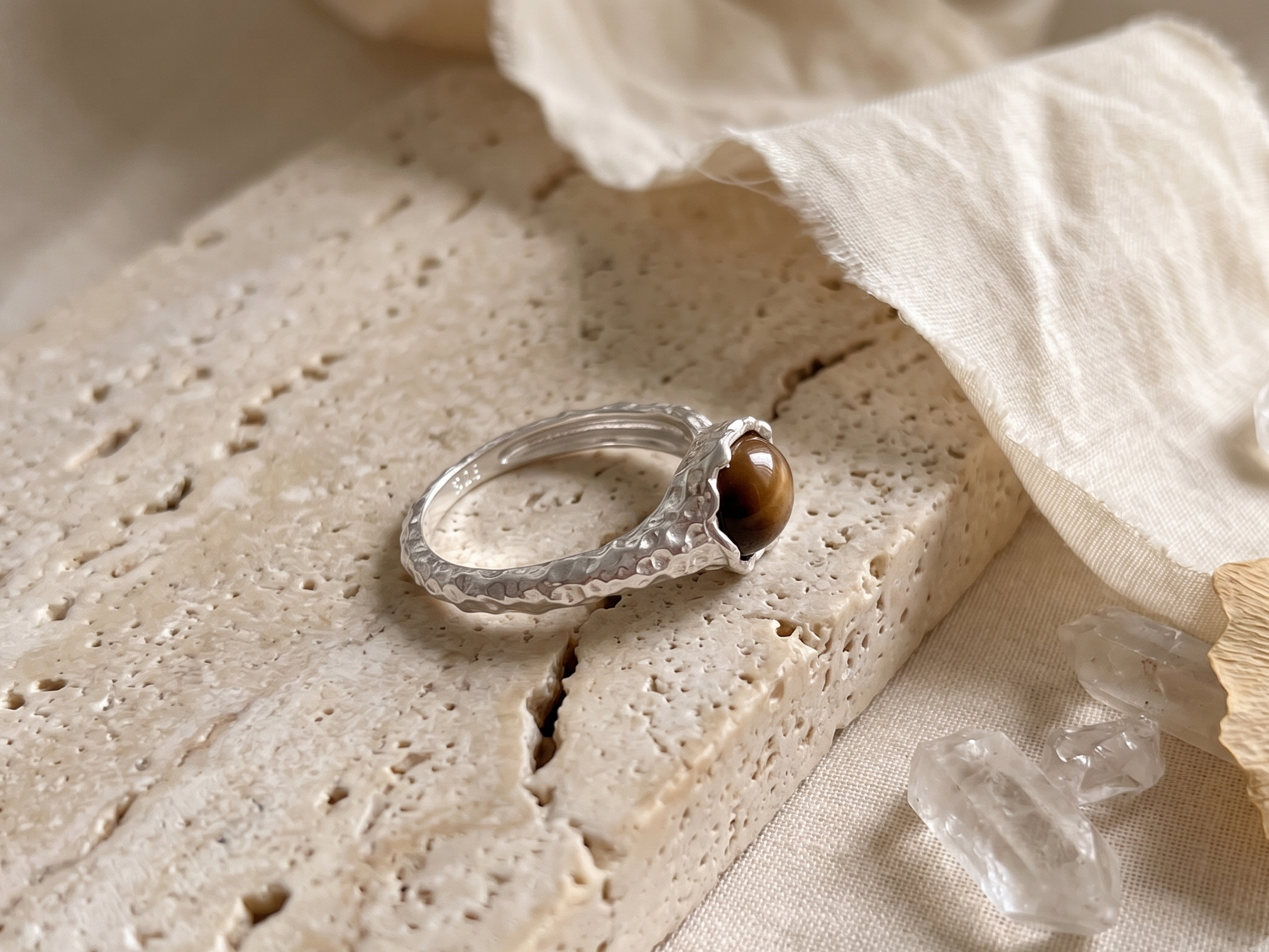 Silver ring with a tiger's eye stone on a textured beige surface with crystals and fabric.