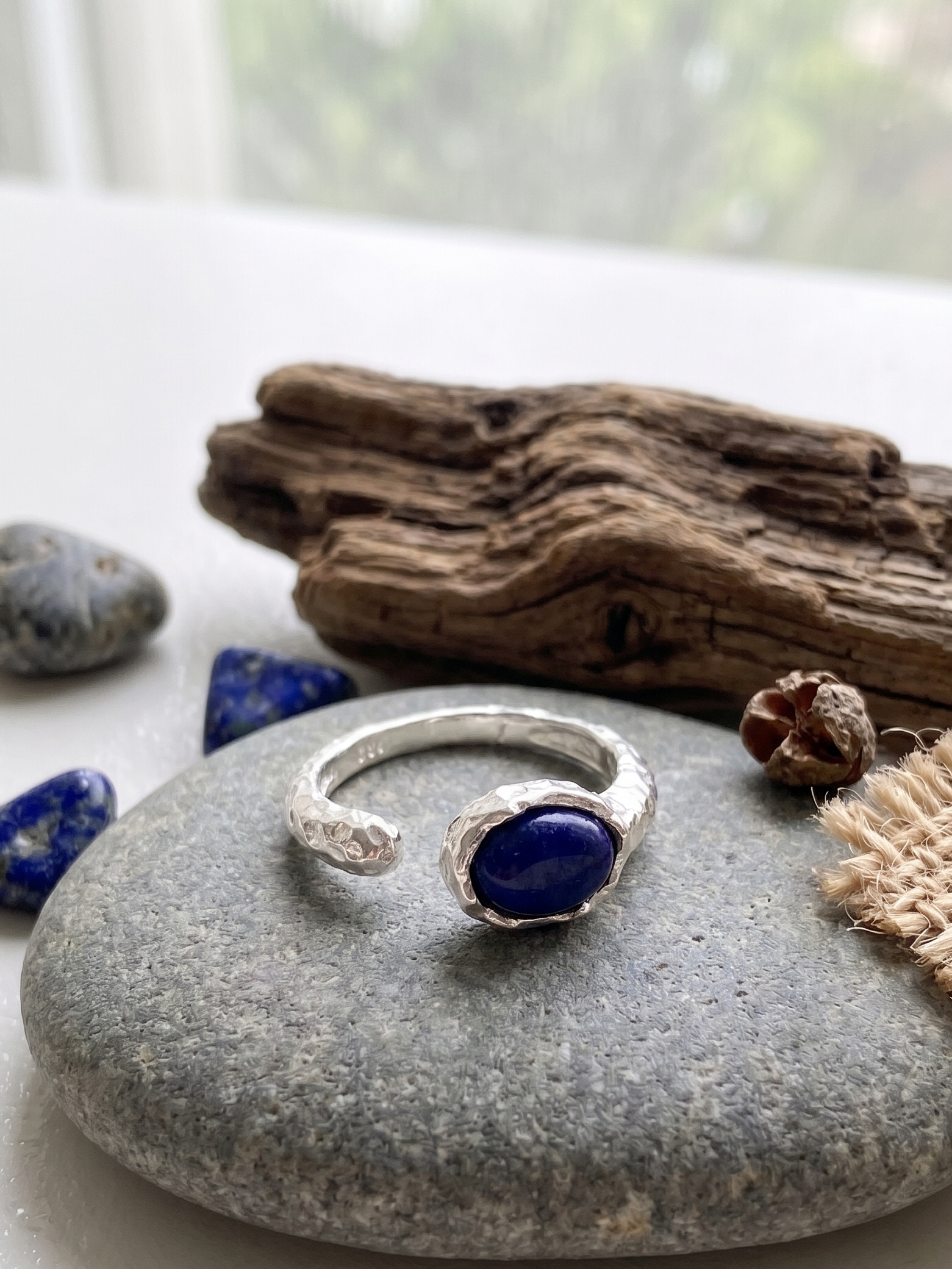 Silver ring with a blue lapis lazuli stone on a gray stone surface with natural elements.