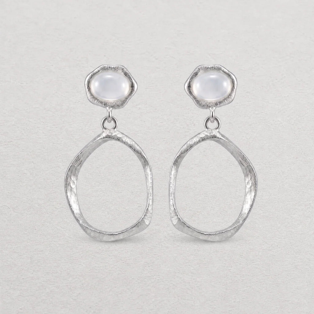 A pair of silver dangle earrings with textured oval drop shapes and smooth, dome-shaped white agate stones at the top, displayed on a light textured background.