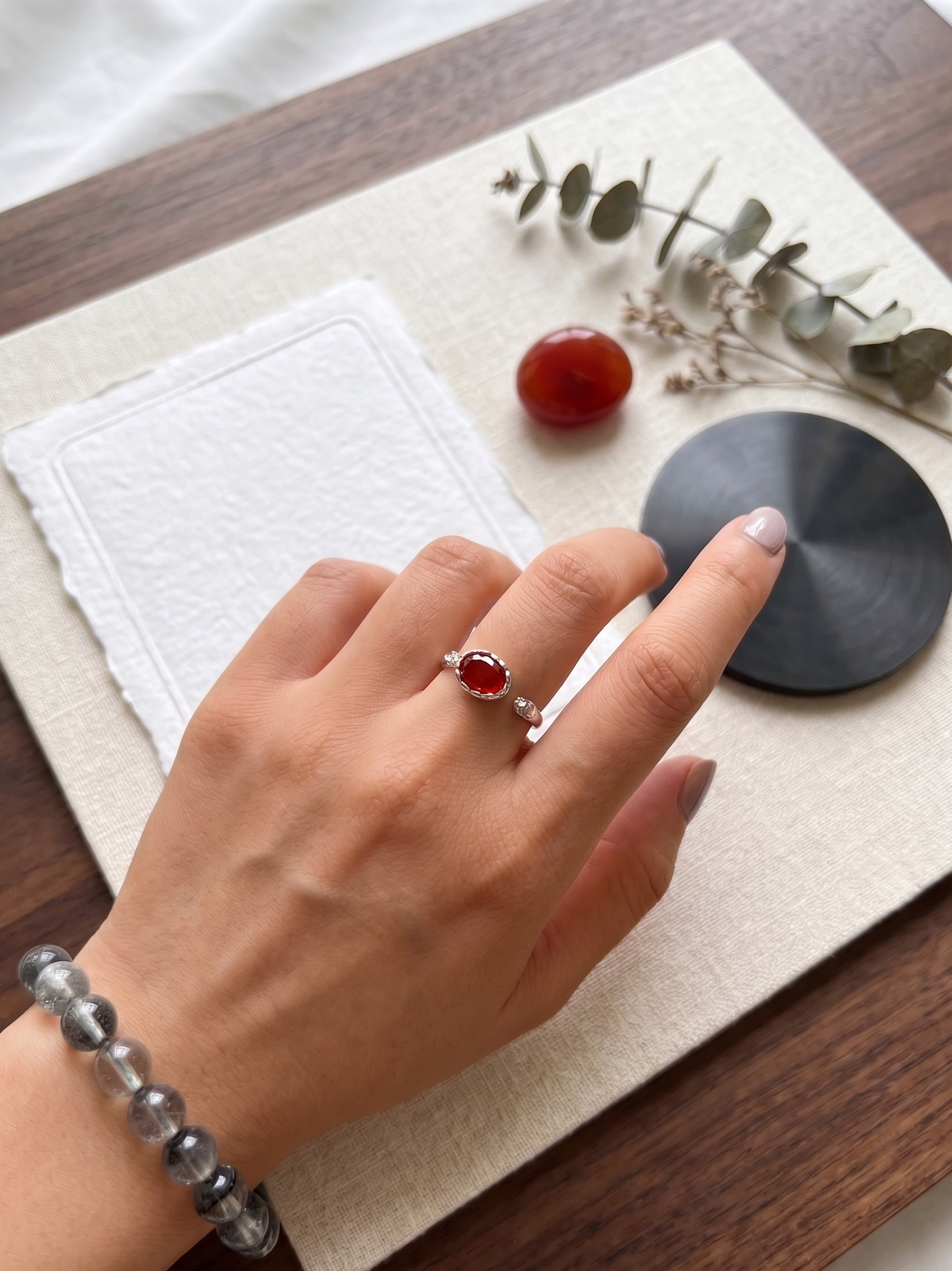 Hand wearing a ring with a red agate stone on a wooden surface with a white cloth and small plant.