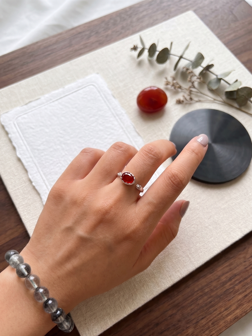 Hand wearing a ring with a red agate stone on a wooden surface with a white cloth and small plant.